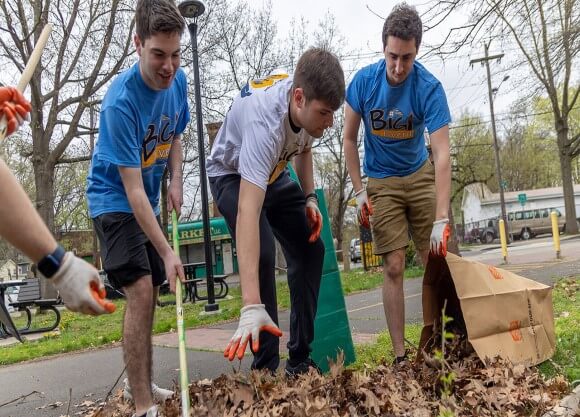 three smiling students rake leaves into a paper bag, grabbing fist fulls of the leaves