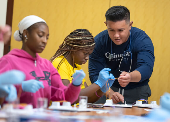 Two high school students wear surgical gloves and work with a faculty member on a health sciences skill