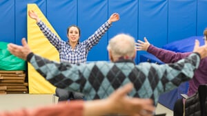 A student raises her arms in front of several people also raising their arms to exercise