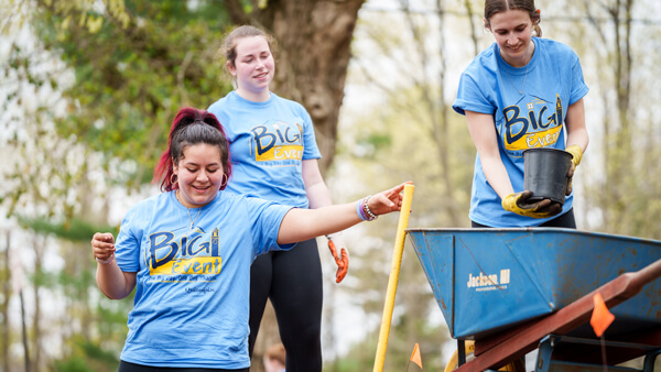 Three students in Quinnipiac Big Event tee shirts work with rakes and a wheelbarrow