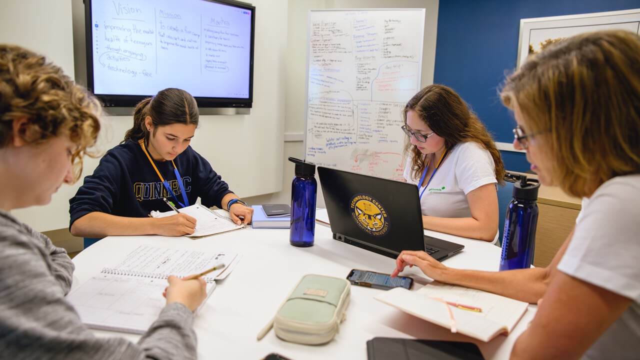 Girl Scouts and leaders sit around a table collaborating with computers and white boards