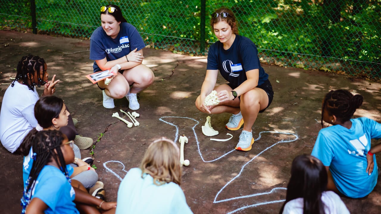 Two physical therapy students talk with children seated on the ground