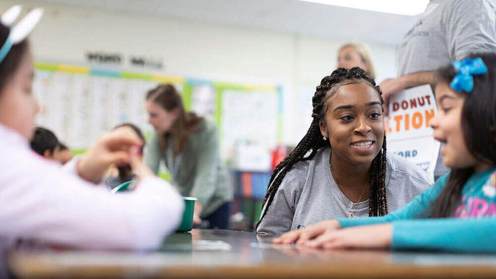 A business student crouches down to interact with second graders seated at their desks